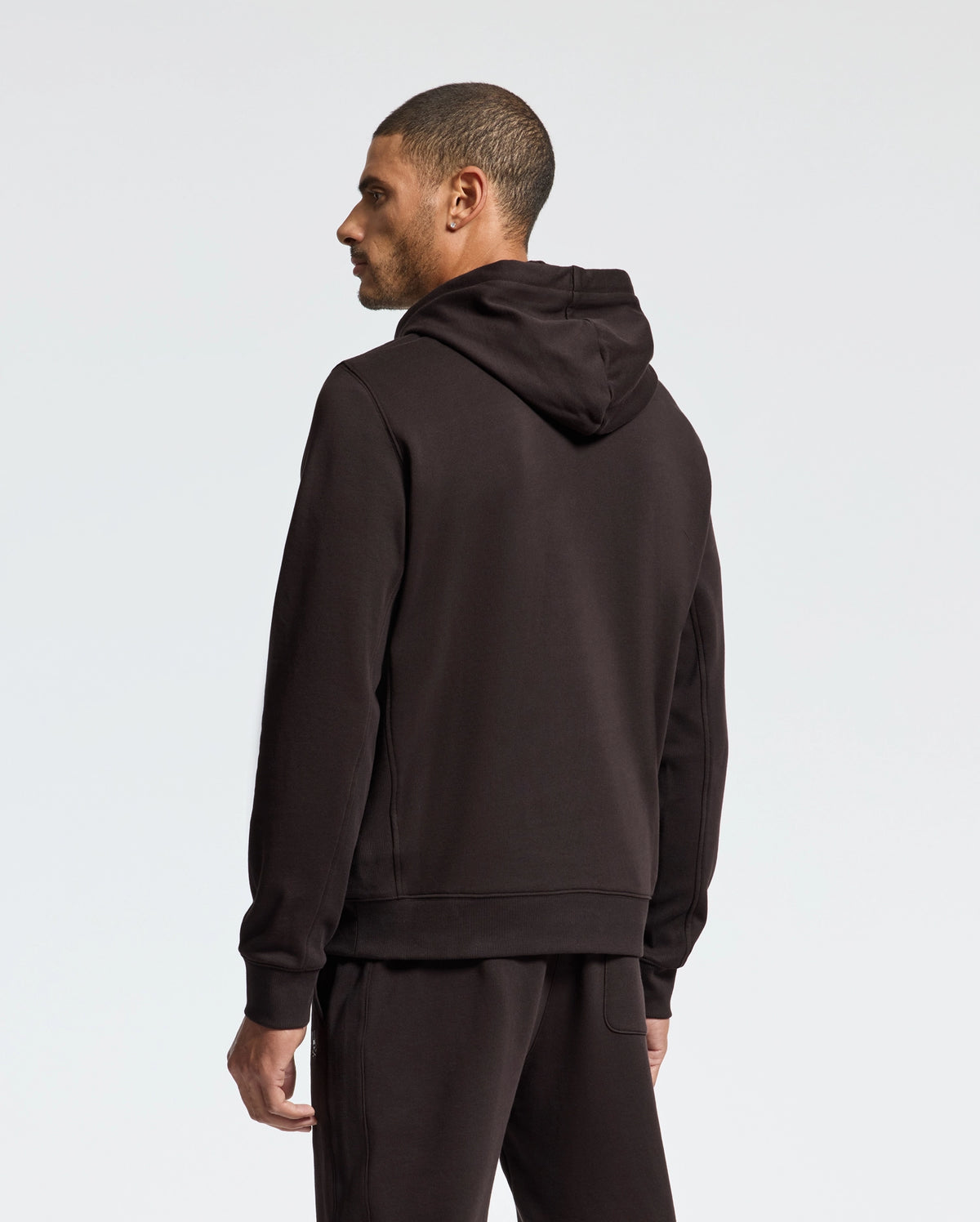 A man in a black Psycho Bunny MENS LEVY ESSENTIAL FLEECE HOODIE - B6H859D200 and matching pants stands, slightly turned to the side. Made from 100% cotton French terry, the outfit's dark tone is accentuated against a plain, light-colored background.