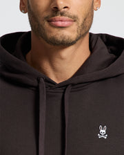 A person is wearing a Psycho Bunny Men's Levy Essential Fleece Hoodie in black, made from 100% cotton French terry. The hoodie is adorned with a small white rabbit and crossbones logo on the chest. The image emphasizes the upper part of the hoodie, highlighting the person's jawline with short facial hair.