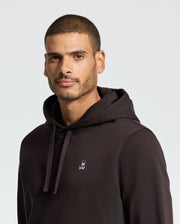 A man with short hair and a light beard is wearing a Psycho Bunny MENS LEVY ESSENTIAL FLEECE HOODIE - B6H859D200, made from 100% cotton French terry and featuring an embroidered logo of a bunny and crossbones. He has a calm expression and is standing against a plain light background.