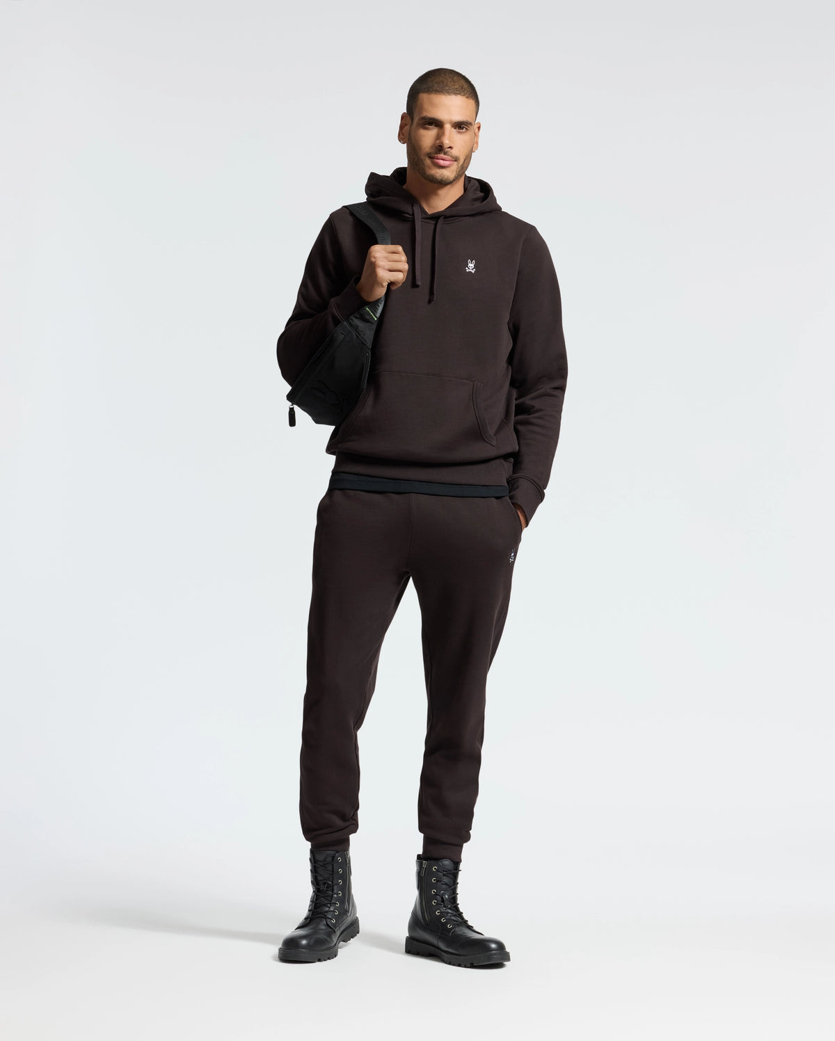 A man is posing in a Psycho Bunny MENS LEVY ESSENTIAL FLEECE HOODIE and joggers set, crafted from soft French terry. He's carrying a black bag over his shoulder and wearing black lace-up boots. The background is plain and light-colored.
