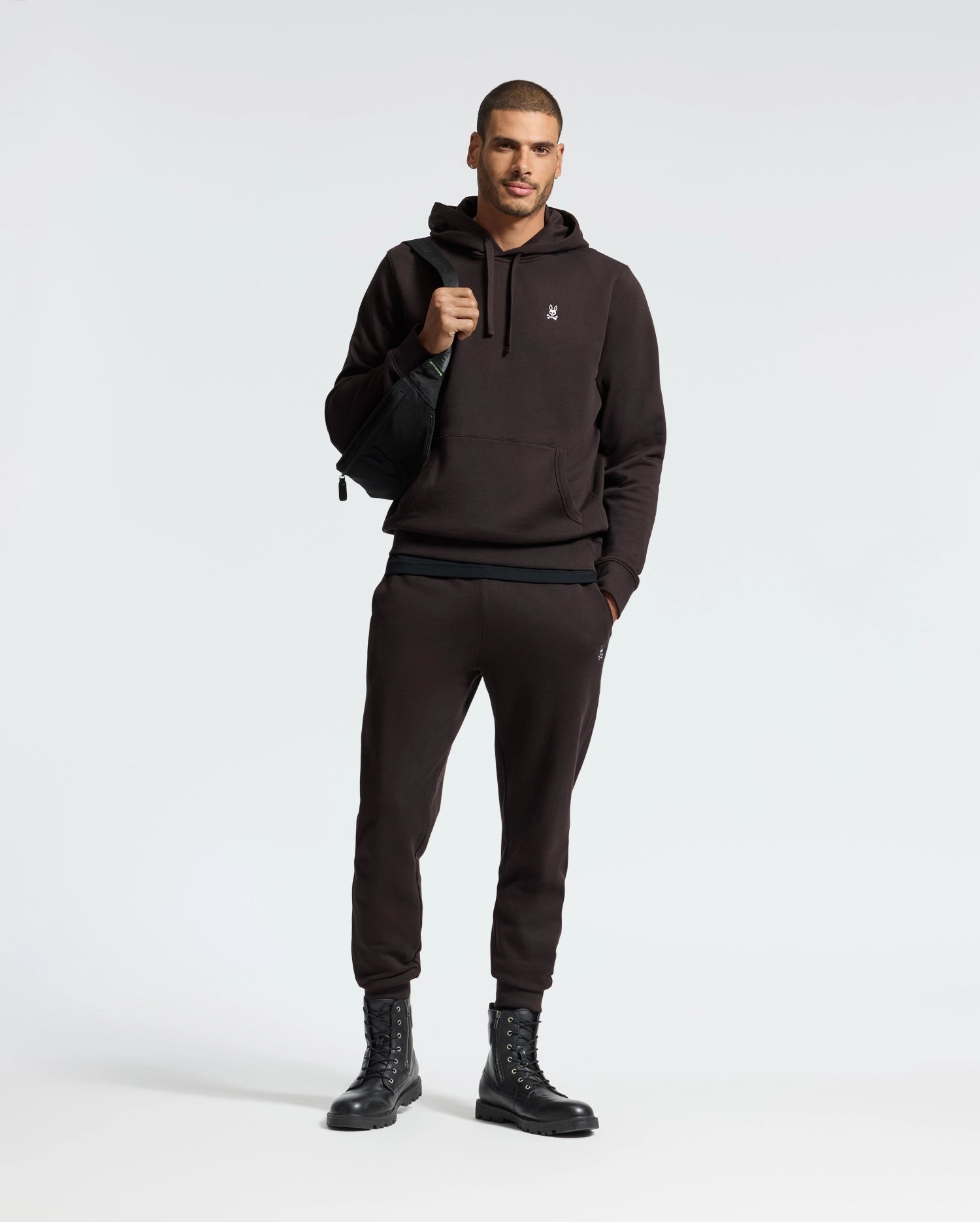 A man is posing in a Psycho Bunny MENS LEVY ESSENTIAL FLEECE HOODIE and joggers set, crafted from soft French terry. He's carrying a black bag over his shoulder and wearing black lace-up boots. The background is plain and light-colored.