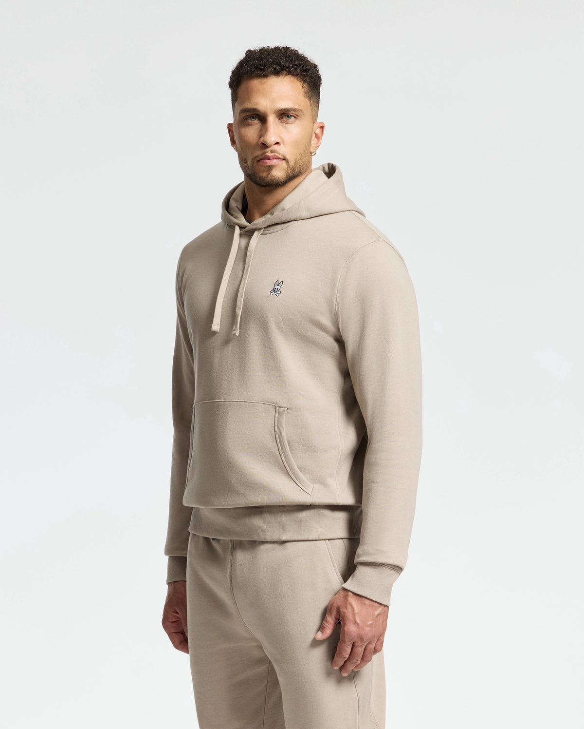 A man wearing the MENS LEVY ESSENTIAL FLEECE HOODIE in beige, by Psycho Bunny, along with matching joggers stands against a plain background. The hoodie displays a small logo on the chest, and he gazes directly at the camera with a neutral expression.