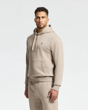A man wearing the MENS LEVY ESSENTIAL FLEECE HOODIE in beige, by Psycho Bunny, along with matching joggers stands against a plain background. The hoodie displays a small logo on the chest, and he gazes directly at the camera with a neutral expression.