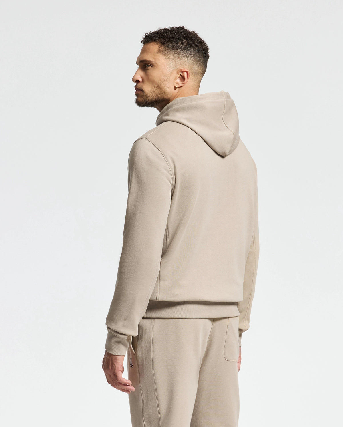 A person wearing the Psycho Bunny MENS LEVY ESSENTIAL FLEECE HOODIE (B6H859D200) and matching sweatpants stands against a plain white background, facing to the left with a focused expression. The fleece hoodie enhances the comfortable and casual vibe of the outfit.