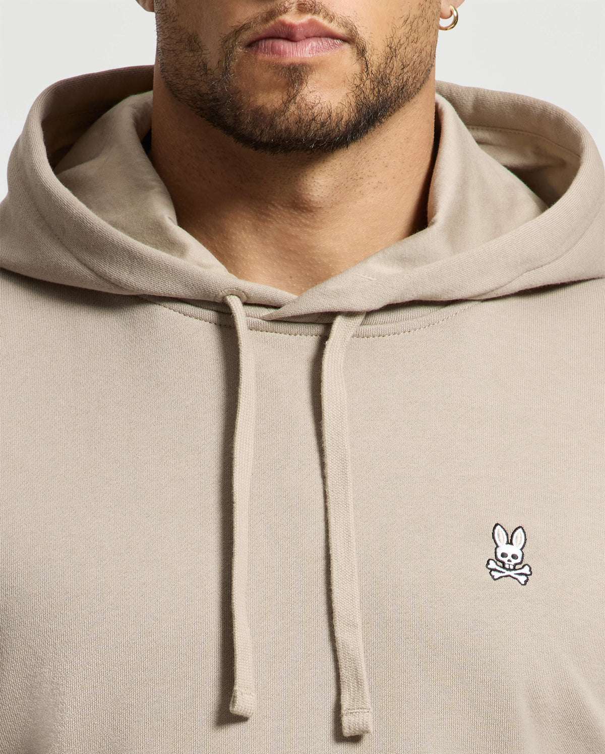 A man wearing the MENS LEVY ESSENTIAL FLEECE HOODIE by Psycho Bunny in beige cotton showcases a small embroidered bunny with crossbones logo on the chest, with the image focusing on his upper body and emphasizing the hoodie's design.
