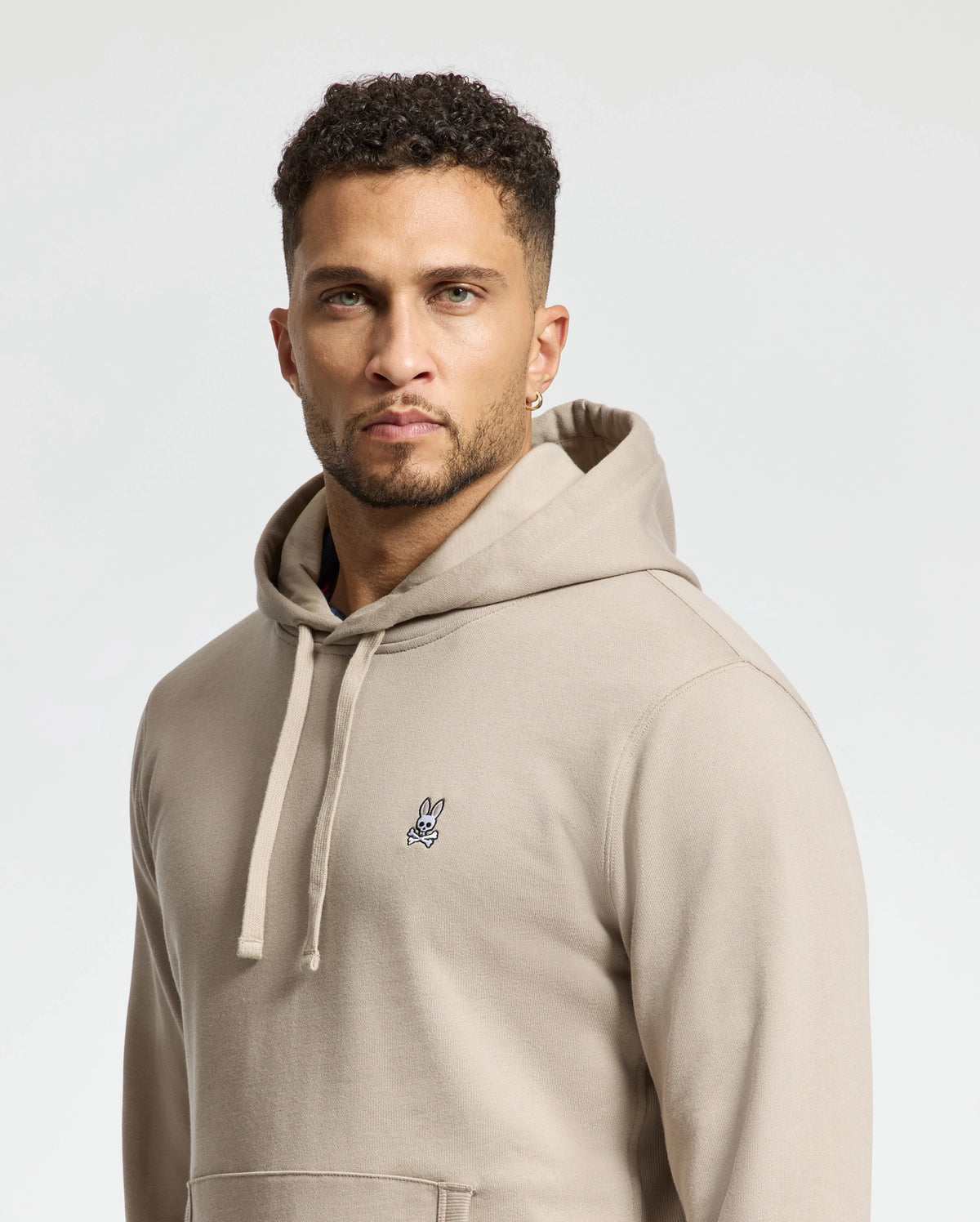A man with short curly hair and a beard is wearing a Psycho Bunny MENS LEVY ESSENTIAL FLEECE HOODIE in beige, featuring a small embroidered design on the chest. The background is plain and light-colored.