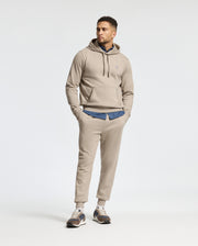 A man wearing the Psycho Bunny MENS LEVY ESSENTIAL FLEECE HOODIE in beige and jogging pants stands with one hand in his pocket. Underneath, he sports a blue shirt and casual sneakers, all set against a plain white background.