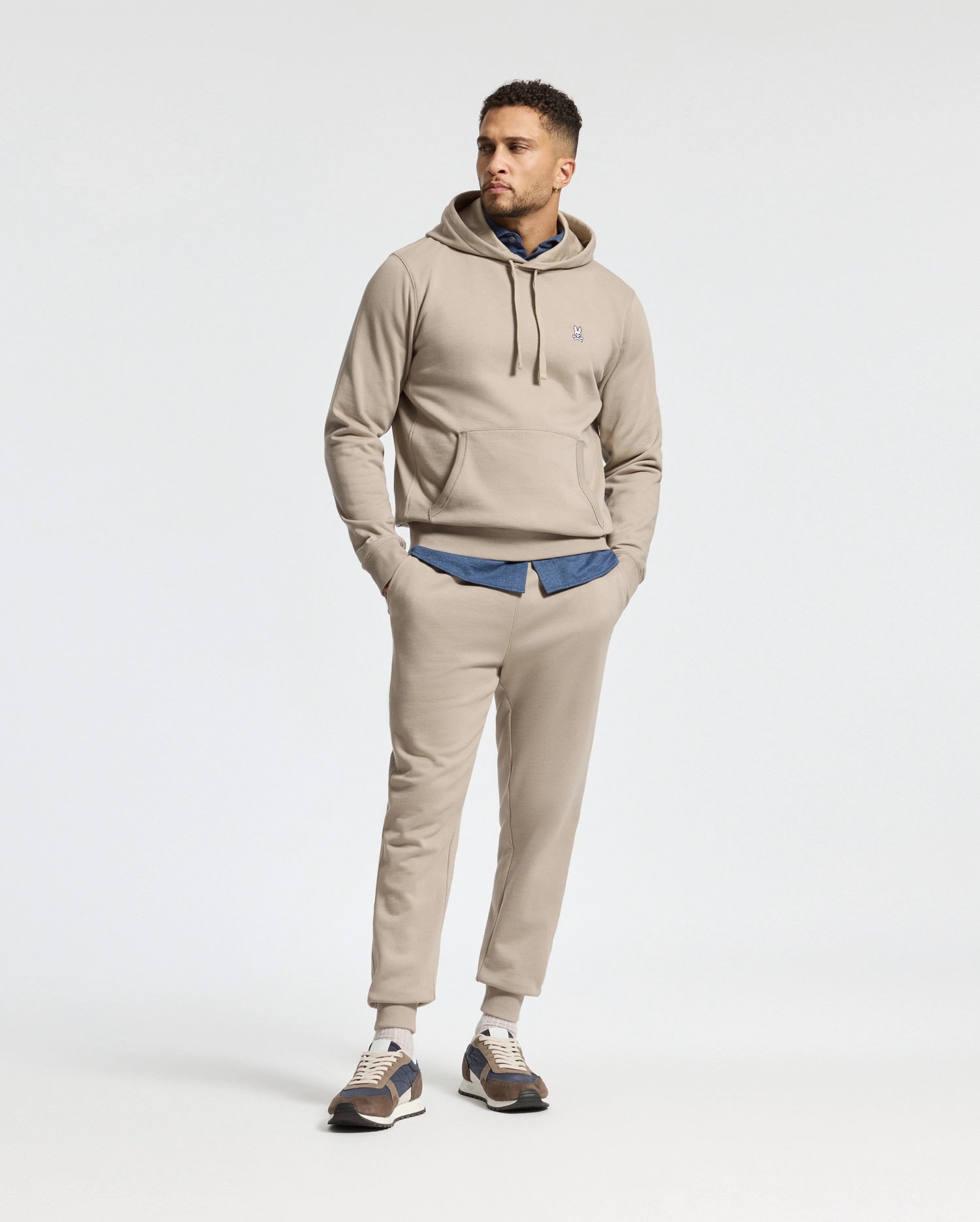 A man wearing the Psycho Bunny MENS LEVY ESSENTIAL FLEECE HOODIE in beige and jogging pants stands with one hand in his pocket. Underneath, he sports a blue shirt and casual sneakers, all set against a plain white background.
