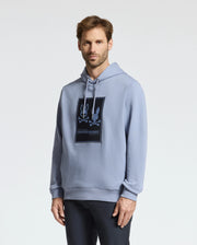 A person wearing a light blue Monroe Twill Patch Fleece Hoodie by Psycho Bunny stands against a plain white background. Made from soft French terry, they have short brown hair and are looking at the camera.