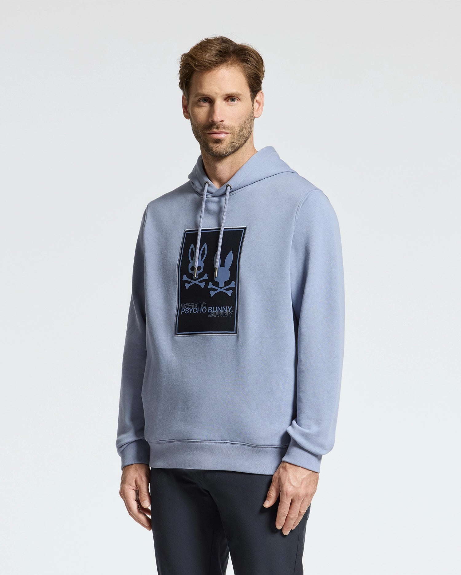 A person wearing a light blue Monroe Twill Patch Fleece Hoodie by Psycho Bunny stands against a plain white background. Made from soft French terry, they have short brown hair and are looking at the camera.