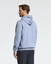 A person stands sideways wearing a light blue Psycho Bunny Monroe Twill Patch Fleece Hoodie and dark pants against a plain background.