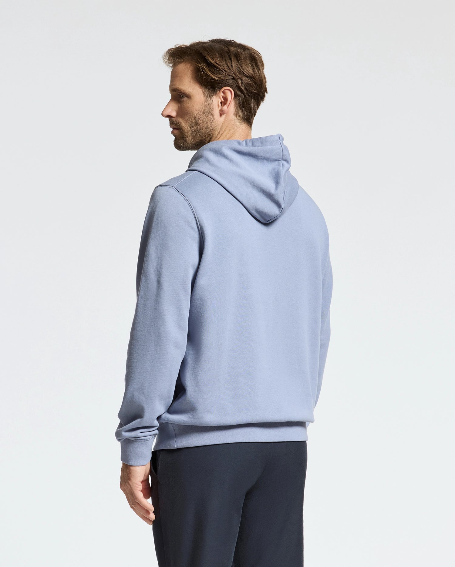 A person stands sideways wearing a light blue Psycho Bunny Monroe Twill Patch Fleece Hoodie and dark pants against a plain background.