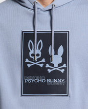 A close-up of the MENS MONROE TWILL PATCH FLEECE HOODIE - B6H926D200 in light blue French terry highlights a graphic design featuring two stylized bunny faces over crossed bones, reminiscent of a skull and crossbones. The brand name "Psycho Bunny" is printed below, emphasizing the distinctive logo appliqué.