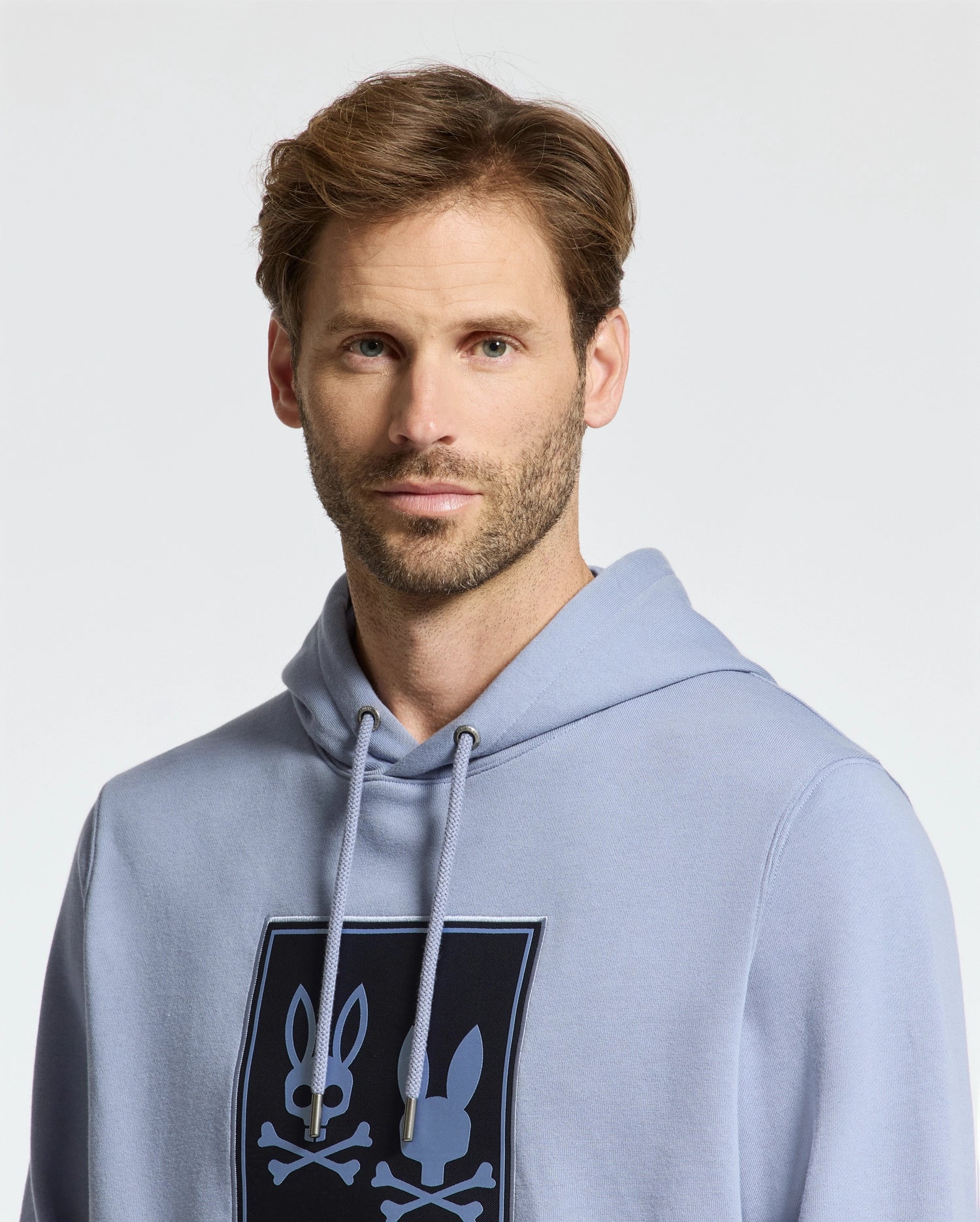 A man with short brown hair and a beard is wearing a Psycho Bunny Monroe Twill Patch Fleece Hoodie in light blue French terry, featuring a black rectangular graphic with two bunny silhouettes and crossbones. The background is plain and light gray.