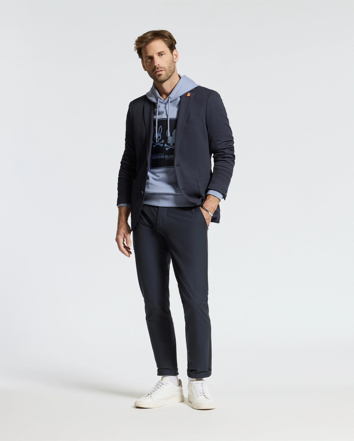 A man stands wearing a coordinated outfit featuring a navy blazer, navy pants, and white sneakers. The ensemble is highlighted by the Psycho Bunny Men's Monroe Twill Patch Fleece Hoodie (B6H926D200), crafted from soft French terry. The plain white background accentuates the smart-casual style, with subtle logo appliqué details adding a touch of sophistication.