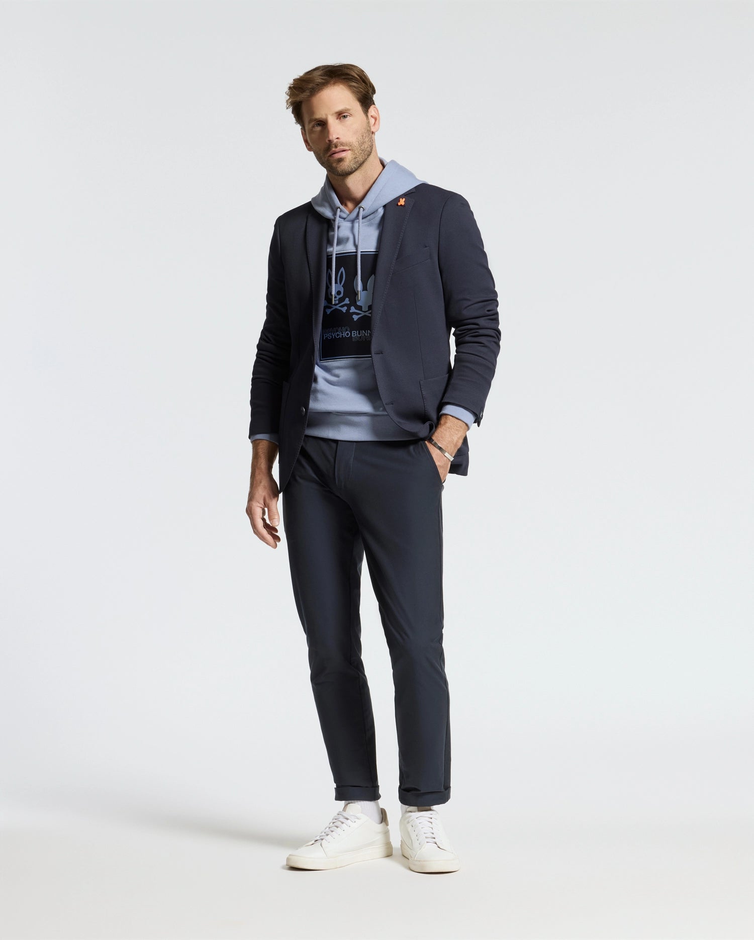 A man stands wearing a coordinated outfit featuring a navy blazer, navy pants, and white sneakers. The ensemble is highlighted by the Psycho Bunny Men's Monroe Twill Patch Fleece Hoodie (B6H926D200), crafted from soft French terry. The plain white background accentuates the smart-casual style, with subtle logo appliqué details adding a touch of sophistication.