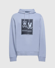 The Psycho Bunny Mens Monroe Twill Patch Fleece Hoodie in light blue features an eye-catching logo appliqué with two stylized rabbits and crossbones, one adorned with a bowtie. Made from French terry fabric, it includes "Psycho Bunny" text along with a hood and long sleeves for enhanced comfort.