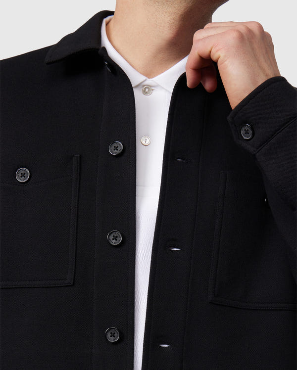 A person is wearing the MENS BEDFORD SHIRT JACKET - B6J290B200 by Psycho Bunny, designed in a work-jacket style with black buttons and visible pockets, over a white collared shirt. The person's hand is adjusting the collar of the shirt, and the image highlights their upper torso.