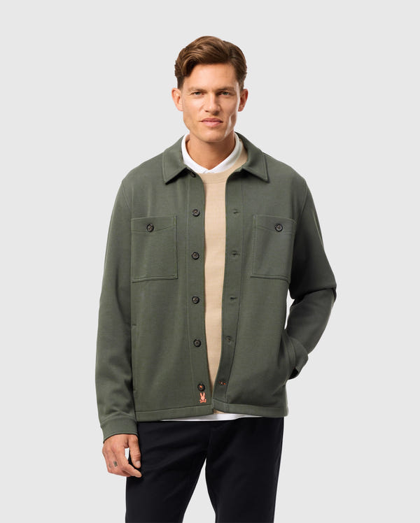 A person wearing the Psycho Bunny MENS BEDFORD SHIRT JACKET - B6J290B200 in green, over a beige sweater, stands against a plain gray background. The jacket, with its urban appeal, showcases two chest pockets. The individual is smiling slightly with one hand tucked into their pocket.