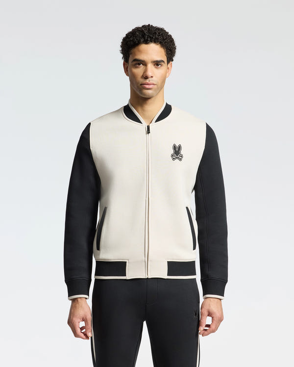 A man showcases the Psycho Bunny MENS CRETE COLOR BLOCK BOMBER (B6J421F200), a beige and black varsity jacket with a classic cut and a subtle embroidered bunny logo on the chest. With short curly hair, he stands against a white background, highlighting this versatile piece perfect for any occasion.