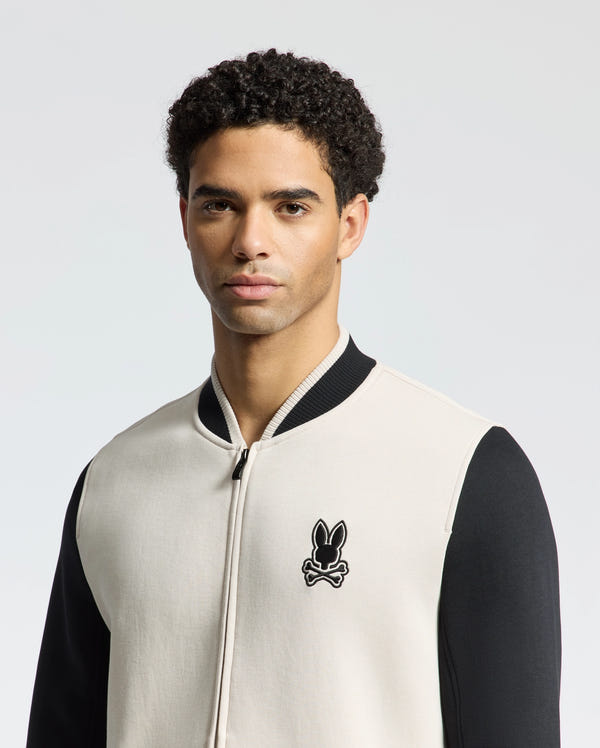 A man with curly hair wears the Psycho Bunny MENS CRETE COLOR BLOCK BOMBER (B6J421F200), a black and white zip-up jacket with a bunny skull logo. His neutral expression enhances the plain light background, making this ensemble an ideal transitional outfit for any occasion.