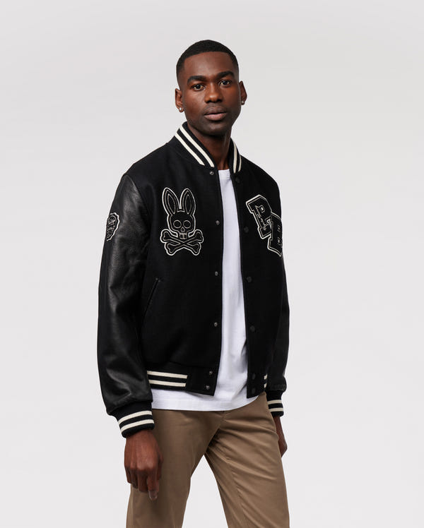 A person wearing the trendy MENS ROBBINS VARSITY BOMBER JACKET - B6J524Z1OW by Psycho Bunny, featuring chenille patches such as a bunny face and the letter 'B'. The wool-blend design pairs seamlessly with their white shirt and khaki pants, set against a simple, minimalistic backdrop.