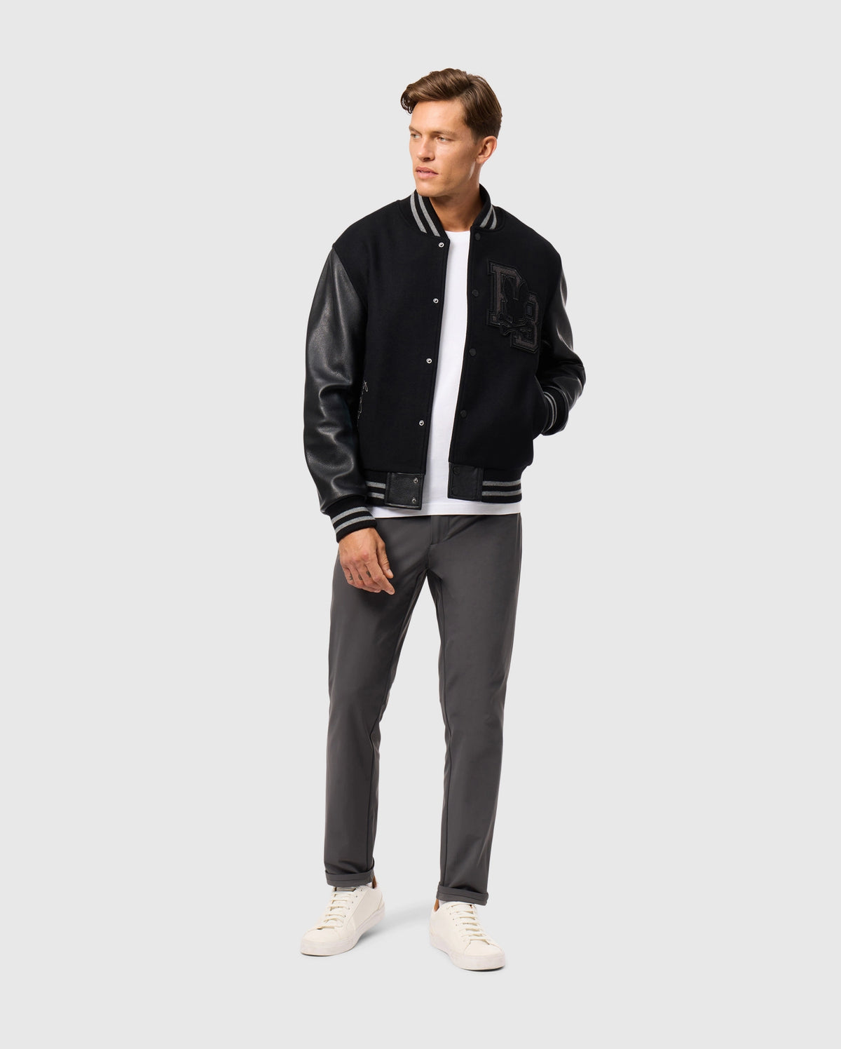 A man stands confidently wearing the Psycho Bunny MENS BRAYDEN WOOL LEATHER VARSITY BOMBER (B6J876D200) with a white t-shirt. He completes his look with gray pants and white sneakers, gazing off to the side against a plain light gray background.