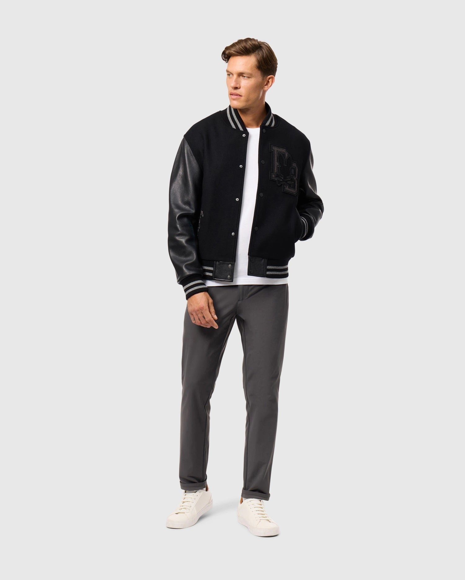 A man stands confidently wearing the Psycho Bunny MENS BRAYDEN WOOL LEATHER VARSITY BOMBER (B6J876D200) with a white t-shirt. He completes his look with gray pants and white sneakers, gazing off to the side against a plain light gray background.