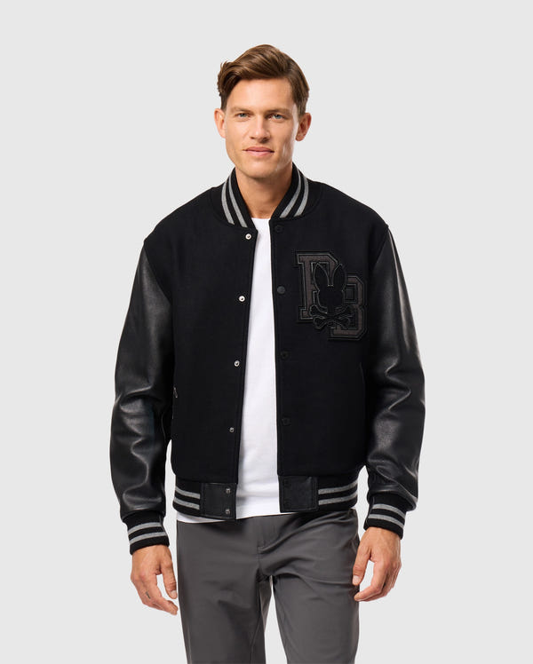 A person is dressed in the MENS BRAYDEN WOOL LEATHER VARSITY BOMBER by Psycho Bunny, featuring leather sleeves and a prominent letter patch on the chest. This stylish wool-blend bomber jacket is paired with a white t-shirt and gray pants, as they stand against a plain gray background.