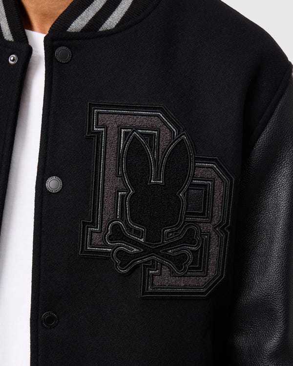 Close-up of the Psycho Bunny MENS BRAYDEN WOOL LEATHER VARSITY BOMBER - B6J876D200, a black bomber-style jacket crafted from a wool blend. It features sleek leather sleeves and is adorned with a prominent "DB" patch displaying a stylized bunny head with crossbones. The jacket also showcases visible button details.