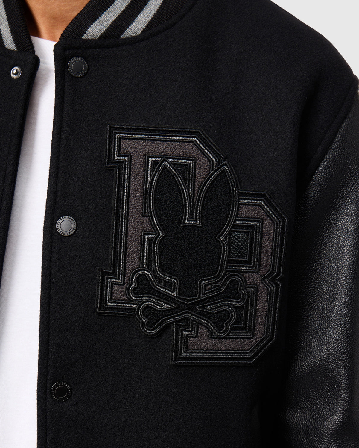 Close-up of the Psycho Bunny MENS BRAYDEN WOOL LEATHER VARSITY BOMBER - B6J876D200, a black bomber-style jacket crafted from a wool blend. It features sleek leather sleeves and is adorned with a prominent "DB" patch displaying a stylized bunny head with crossbones. The jacket also showcases visible button details.