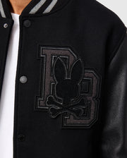 Close-up of the Psycho Bunny MENS BRAYDEN WOOL LEATHER VARSITY BOMBER - B6J876D200, a black bomber-style jacket crafted from a wool blend. It features sleek leather sleeves and is adorned with a prominent "DB" patch displaying a stylized bunny head with crossbones. The jacket also showcases visible button details.