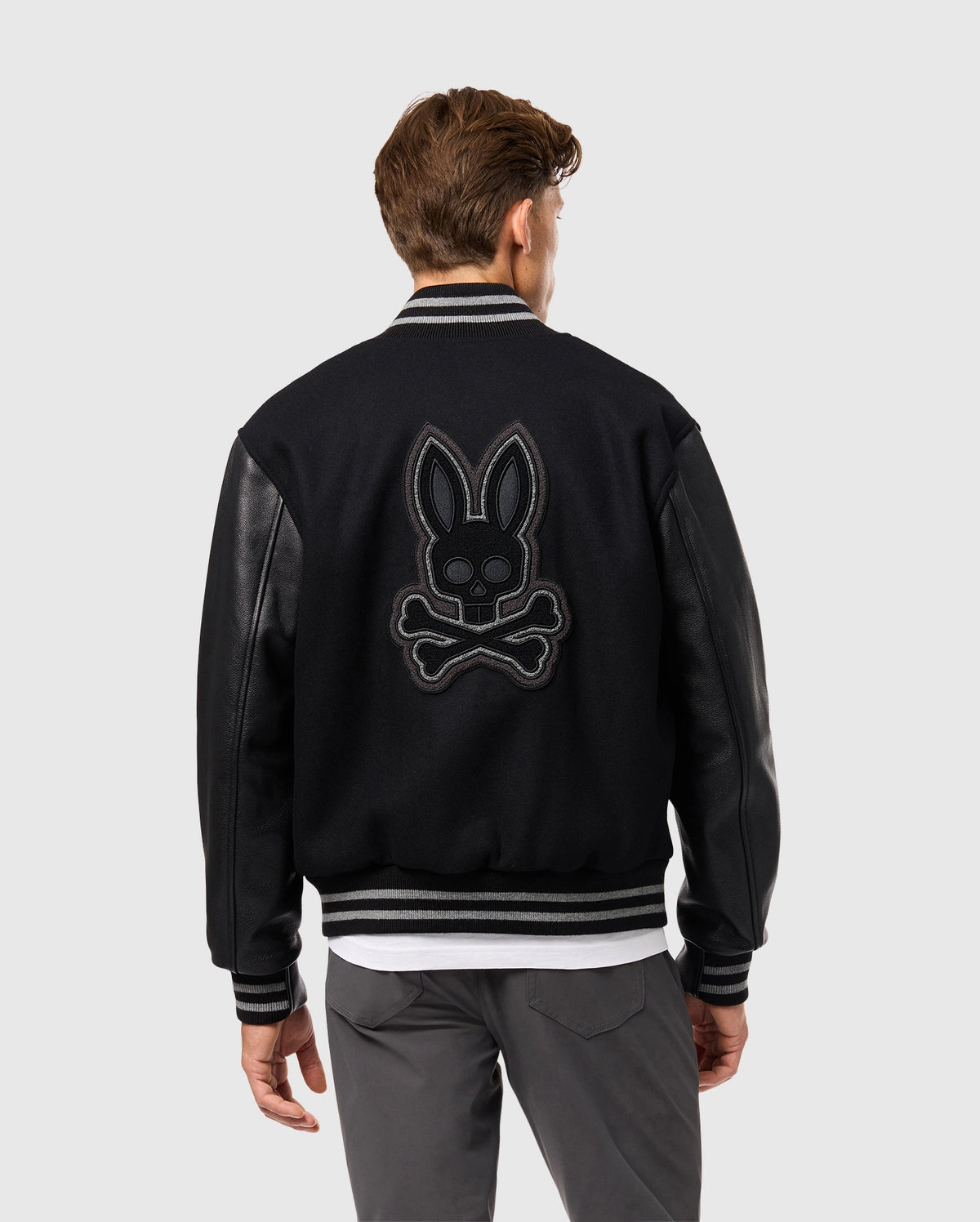 A person is wearing the Psycho Bunny MENS BRAYDEN WOOL LEATHER VARSITY BOMBER - B6J876D200, a black jacket that combines leather sleeves with elements of a wool-blend bomber. The back showcases an embroidered bunny with crossbones design as the individual faces away, drawing attention to the striking detail.