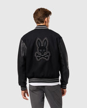 A person is wearing the Psycho Bunny MENS BRAYDEN WOOL LEATHER VARSITY BOMBER - B6J876D200, a black jacket that combines leather sleeves with elements of a wool-blend bomber. The back showcases an embroidered bunny with crossbones design as the individual faces away, drawing attention to the striking detail.
