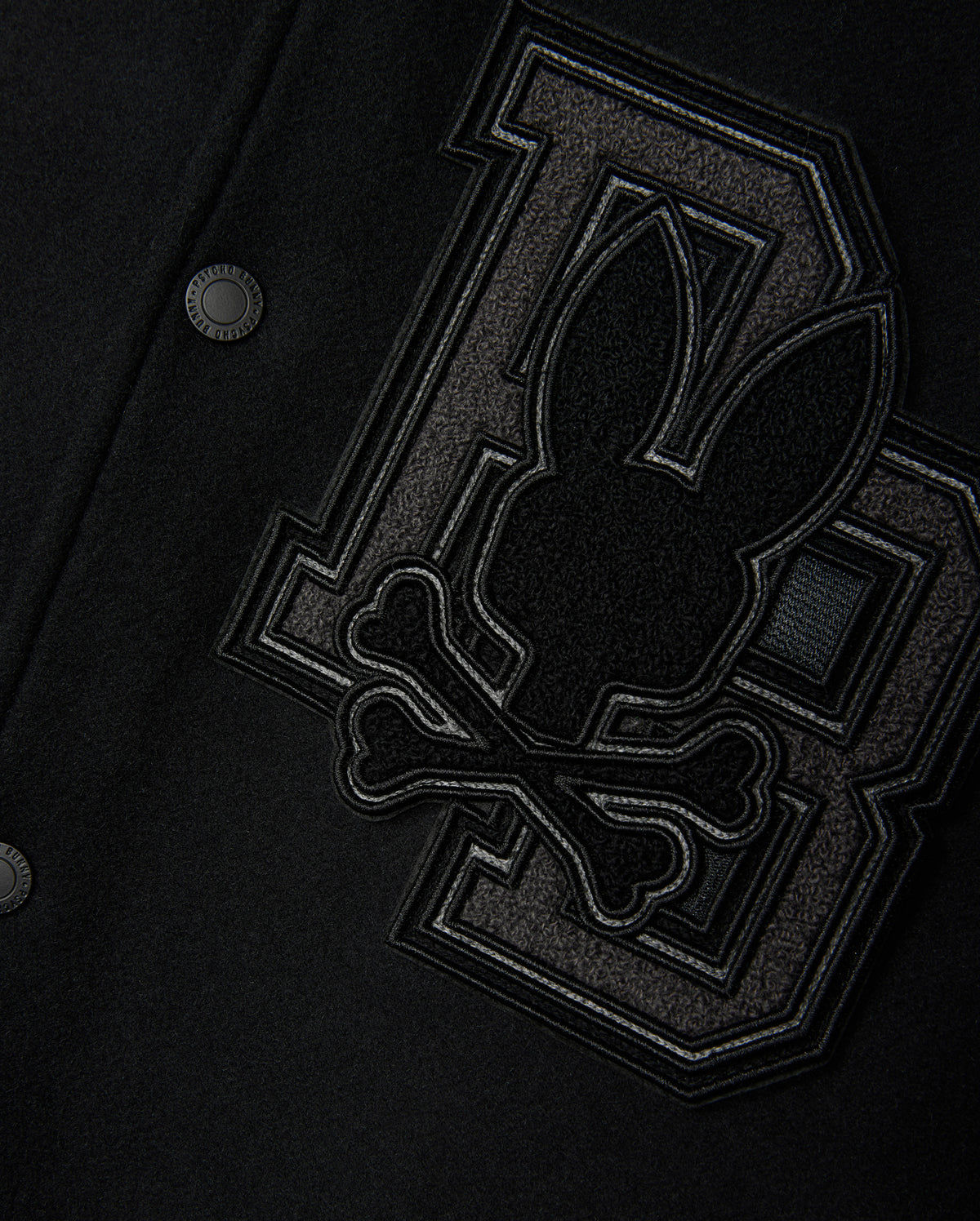 Close-up of the Psycho Bunny MENS BRAYDEN WOOL LEATHER VARSITY BOMBER - B6J876D200, a black wool-blend bomber jacket adorned with large, textured "D" and "B" letters, along with a stitched emblem showcasing a stylized rabbit head and crossbones in dark fabric. The image emphasizes the intricate detailing and craftsmanship characteristic of a classic varsity jacket.