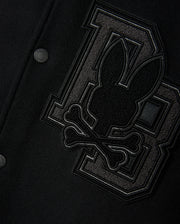 Close-up of the Psycho Bunny MENS BRAYDEN WOOL LEATHER VARSITY BOMBER - B6J876D200, a black wool-blend bomber jacket adorned with large, textured "D" and "B" letters, along with a stitched emblem showcasing a stylized rabbit head and crossbones in dark fabric. The image emphasizes the intricate detailing and craftsmanship characteristic of a classic varsity jacket.