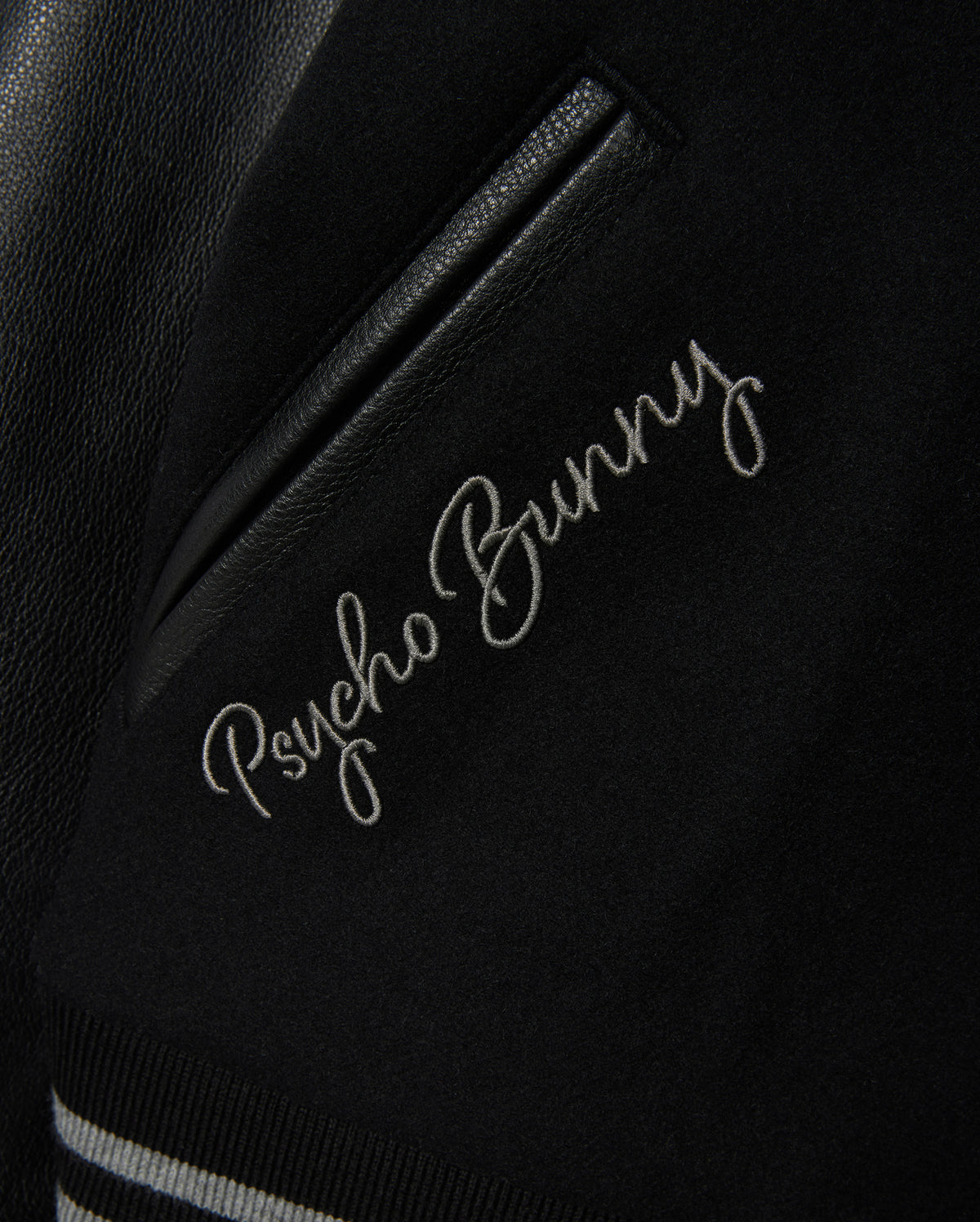 Close-up of the MENS BRAYDEN WOOL LEATHER VARSITY BOMBER jacket, featuring "Psycho Bunny" embroidered near a pocket. This black varsity piece showcases leather sleeves and is enhanced with striped ribbed trimming for a stylish finish.