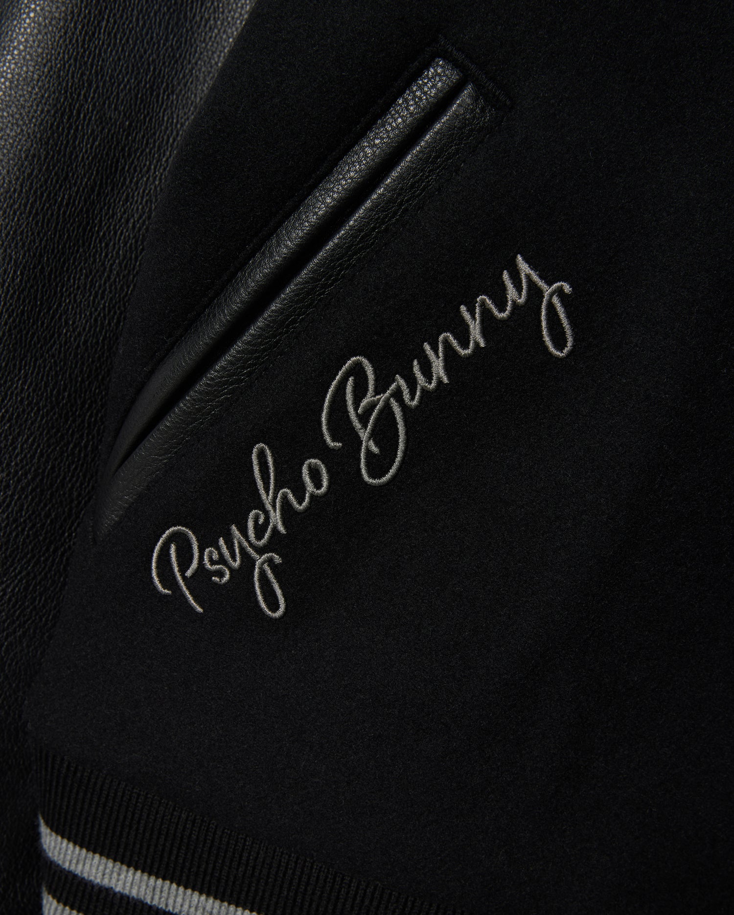 Close-up of the MENS BRAYDEN WOOL LEATHER VARSITY BOMBER jacket, featuring "Psycho Bunny" embroidered near a pocket. This black varsity piece showcases leather sleeves and is enhanced with striped ribbed trimming for a stylish finish.