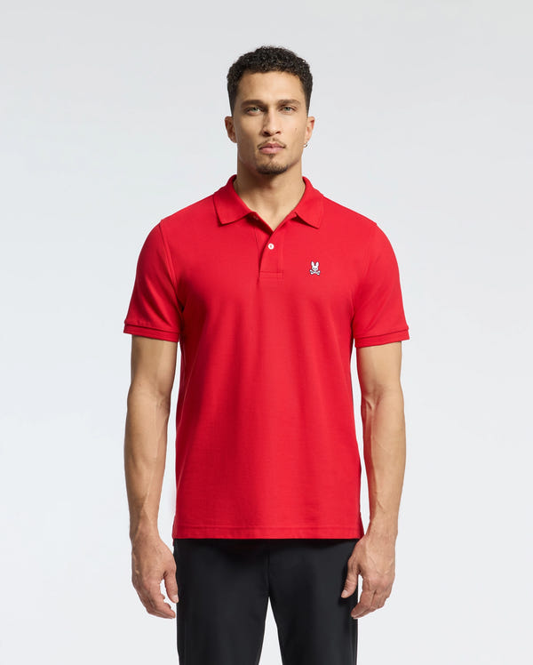 A man stands against a light gray background wearing a vivid red Psycho Bunny Men's Classic Pique Polo Shirt (B6K001CRPC) made from premium Pima cotton. The shirt features a small embroidered logo on the chest and is paired with black pants. His neutral expression complements his short curly hair, while the subtle mother-of-pearl buttons add an elegant touch.