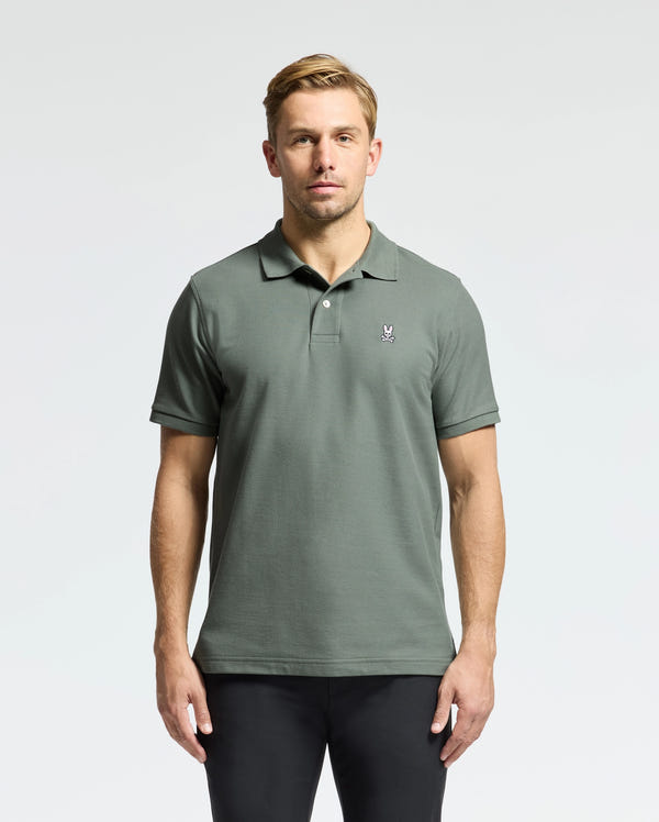 A man in a Psycho Bunny MENS CLASSIC PIQUE POLO SHIRT, featuring an embroidered Bunny logo on the chest, stands against a plain background. He has short blonde hair and is wearing black pants.