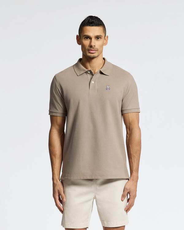 A man models Psycho Bunny's Classic Pique Polo Shirt (B6K001CRPC) in beige Pima cotton, featuring a small chest logo. He's paired it with light beige shorts against a plain white backdrop.