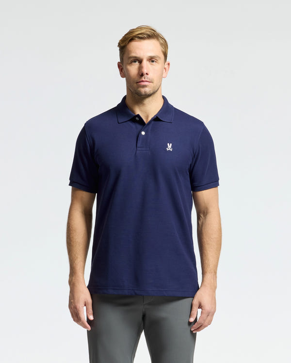 A man with short, blonde hair is wearing the Psycho Bunny MENS CLASSIC PIQUE POLO SHIRT - B6K001CRPC in navy blue along with gray pants. The polo is adorned with a small white logo on the chest. He gazes directly at the camera against a simple, light gray backdrop.