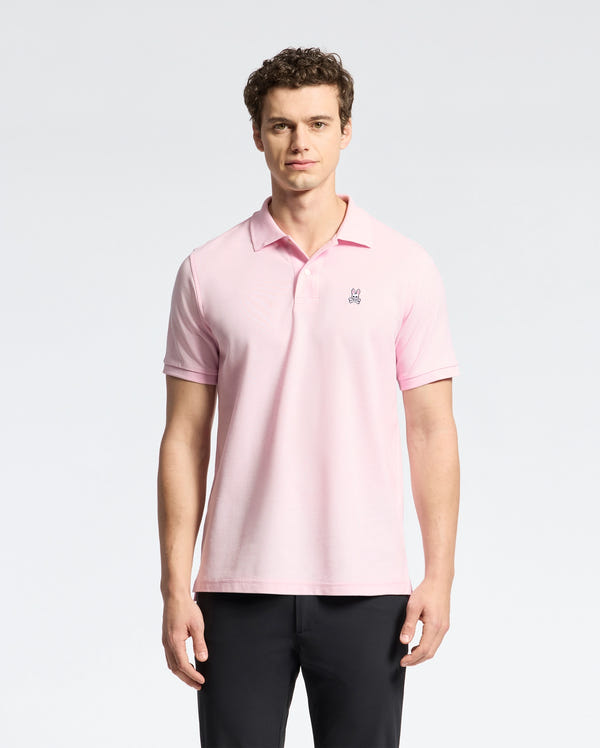 A person dons a Psycho Bunny Men's Classic Pique Polo Shirt - B6K001CRPC in pink, made from Pima cotton with an embroidered chest logo, against a plain light background. Facing the camera neutrally, they pair it with dark pants.