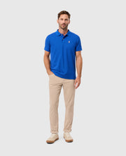 A man wearing a Psycho Bunny MENS CLASSIC PIQUE POLO SHIRT - B6K001CRPC, distinguished by its embroidered Bunny logo, stands against a simple white backdrop. His vibrant blue polo is paired with beige pants and matching sneakers, with one hand nonchalantly tucked in his pocket.