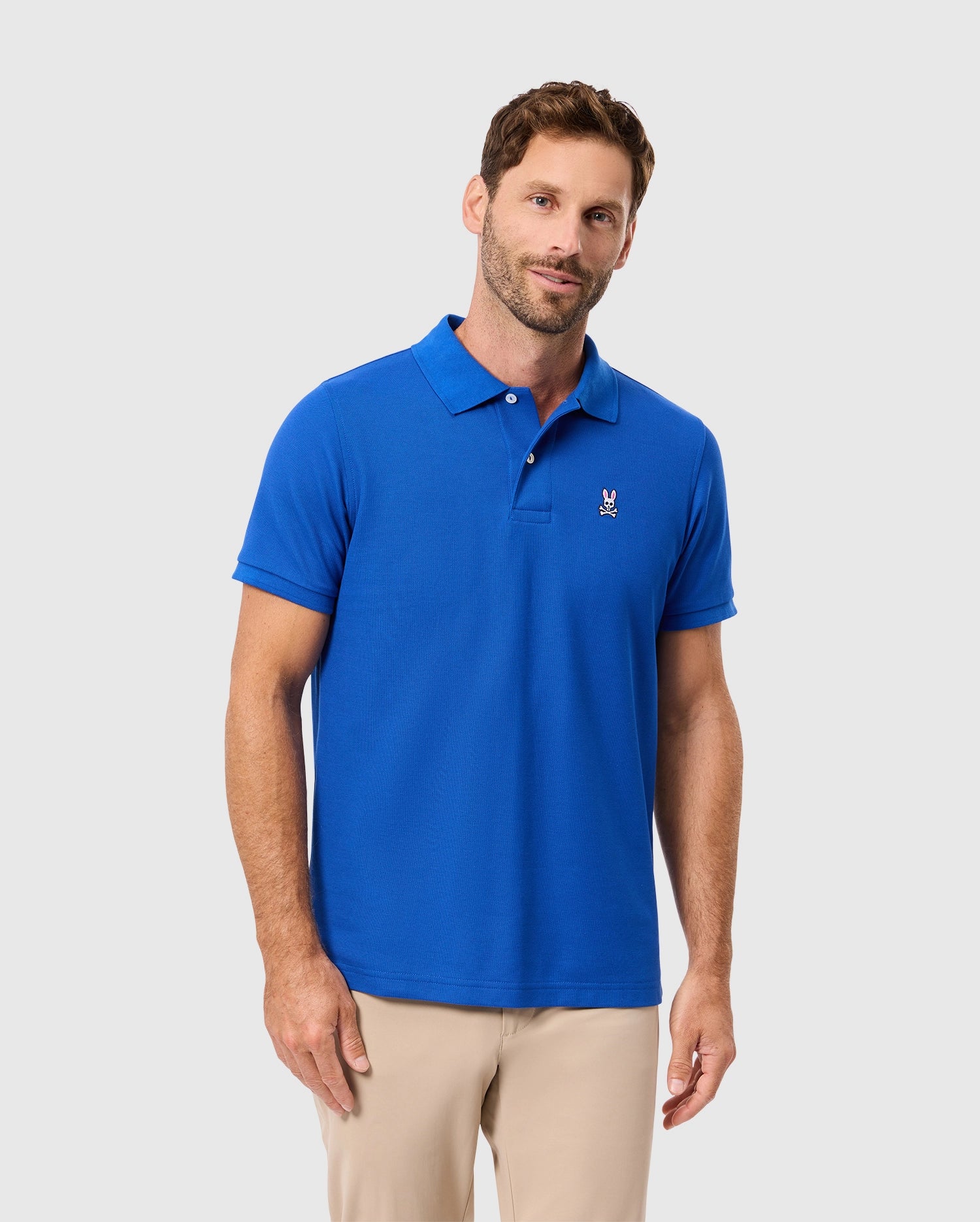 A person dressed in the Psycho Bunny MENS CLASSIC PIQUE POLO SHIRT - B6K001CRPC, featuring an embroidered Bunny logo on the chest in bright blue, paired with beige pants, stands against a plain light gray background. They sport short hair and a beard.