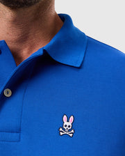 A person wearing a Psycho Bunny MENS CLASSIC PIQUE POLO SHIRT - B6K001CRPC in blue, adorned with the brand's signature embroidered Bunny logo of a rabbit skull and crossbones on the chest.