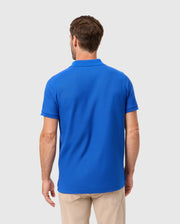 A man with short hair stands with his back to the camera in a bright blue Psycho Bunny MENS CLASSIC PIQUE POLO SHIRT - B6K001CRPC, showcasing an embroidered Bunny logo, paired with beige pants against a plain white background.