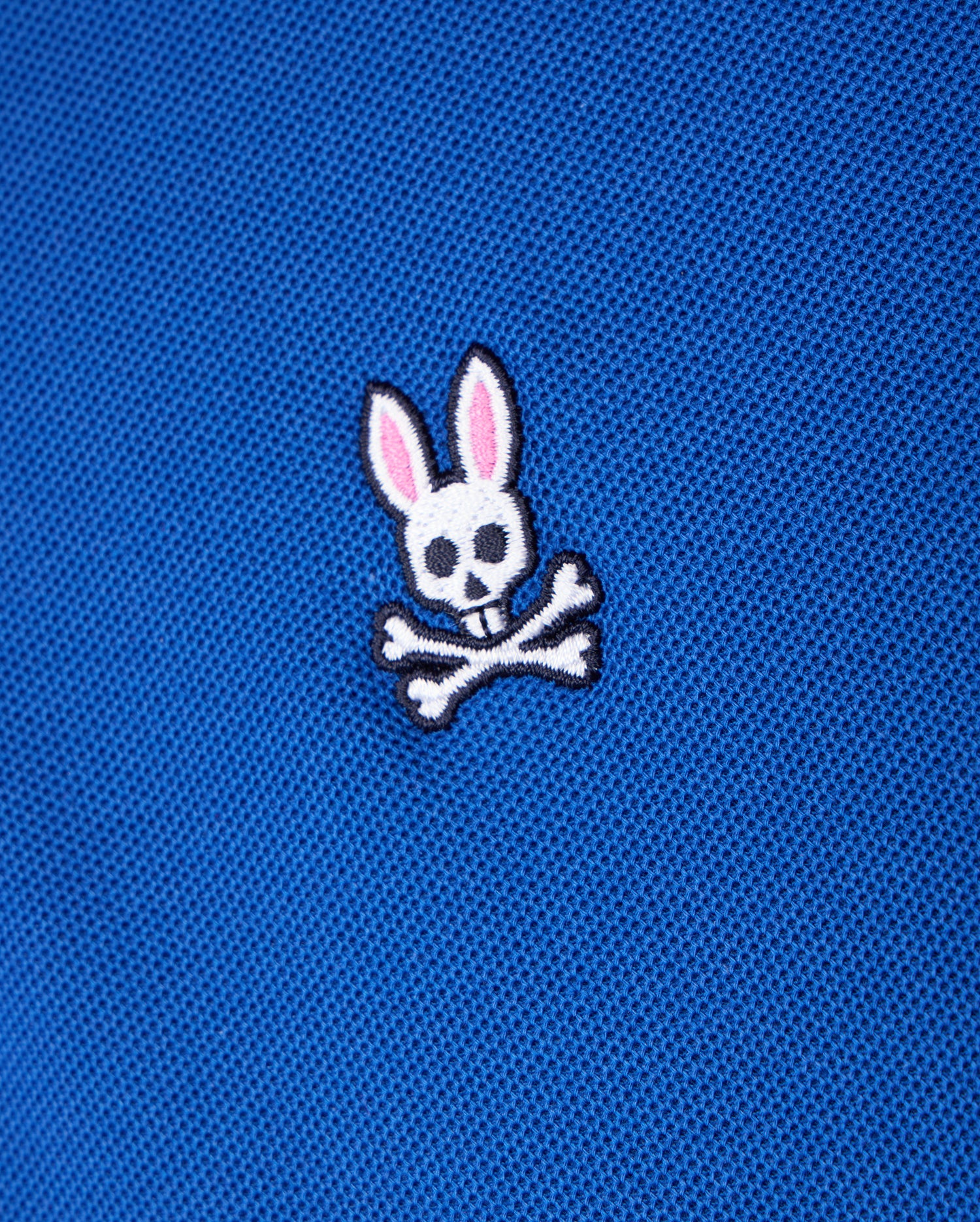 The Psycho Bunny Men's Classic Piqué Polo Shirt - B6K001CRPC features a close-up of blue fabric highlighting the iconic embroidered Bunny logo. This design includes a white skull with rabbit ears and pink inner ears, set above two crossed bones, making it a timeless choice for your luxurious Pima cotton piqué polo.