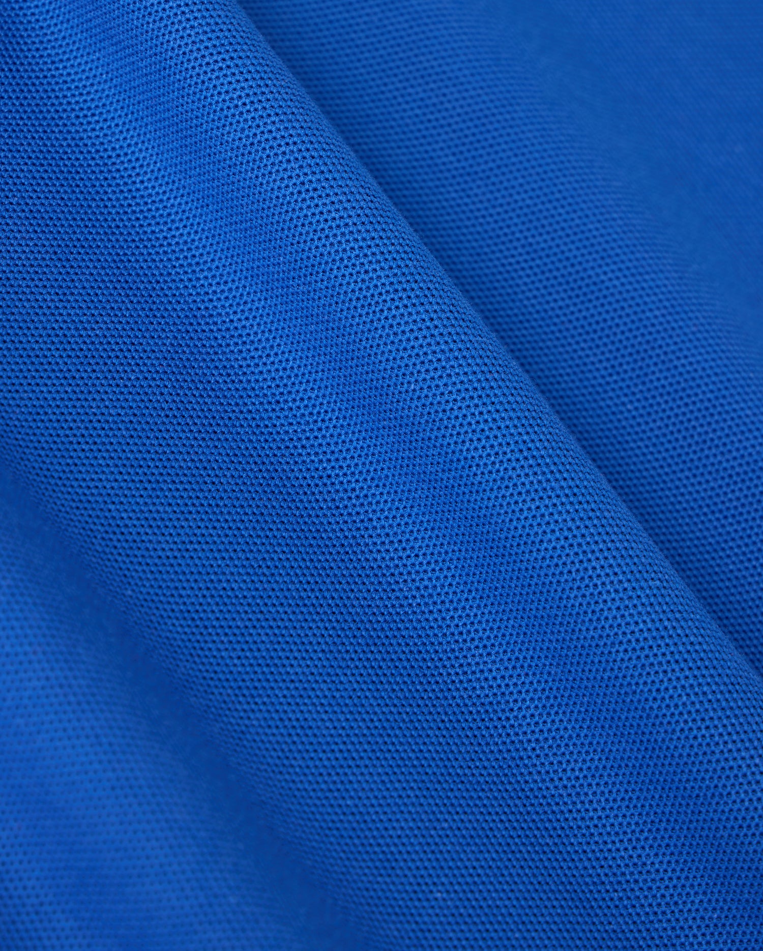 Close-up of blue fabric from the Psycho Bunny MENS CLASSIC PIQUE POLO SHIRT - B6K001CRPC, featuring a classic piqué polo texture. The material is lightweight and semi-transparent with uniformly distributed tiny holes throughout, creating a patterned appearance akin to fine Pima cotton.