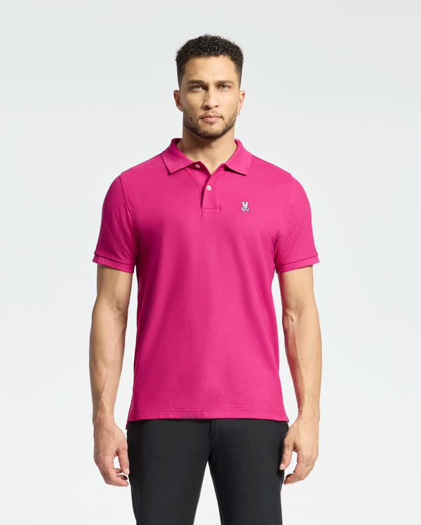 A man against a white background wears a vibrant pink Psycho Bunny MENS CLASSIC PIQUE POLO SHIRT - B6K001CRPC with the logo on the chest. His short curly hair and black pants create an effortlessly stylish look.